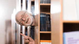Librarian leaning out from behind shelves whilst making a gesture that indicates he has an idea, he looks happy